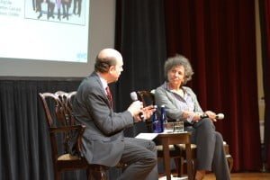 On a stage, a man in a suit holding a microphone interviews a woman.