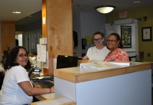 in medical clinic setting, three women pose behind a counter