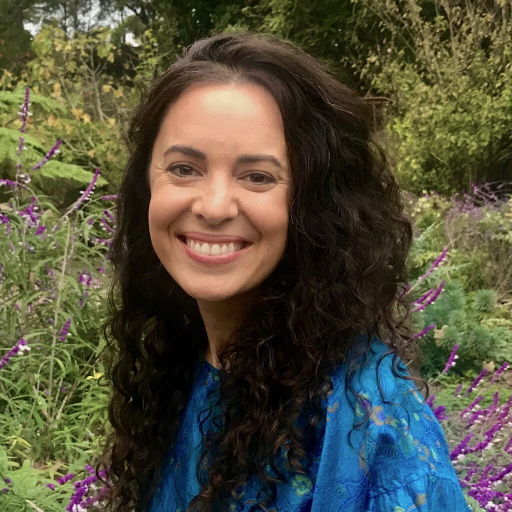 Color photo of a smiling woman with long, curly brown hair. She's wearing a blue flowered blouse, and purple flowers are in the background.