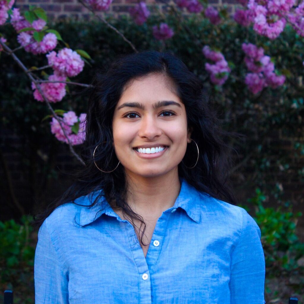 Color photo of a woman smiling. She has long black hair and she's wearing a blue blouse. Pink-purple cherry blossoms are in the background.